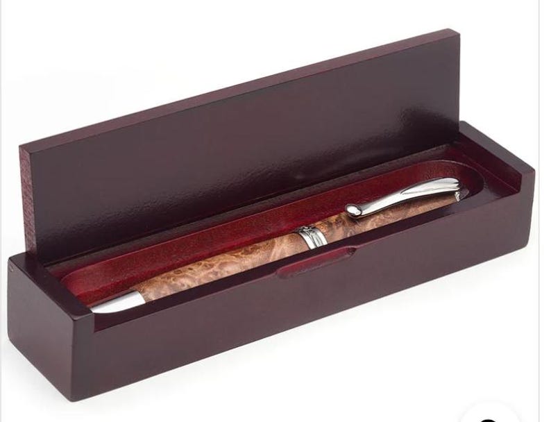May include: A wooden pen set in a dark brown box with a hinged lid. The pen has a silver-colored clip and tip, and a patterned, light brown barrel. The interior of the box is lined with a red fabric.