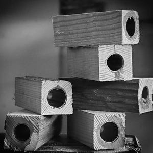 May include: A stack of six wooden blocks with circular metal inserts. The blocks are rectangular and appear to be made of unfinished wood. The metal inserts are visible at the ends of the blocks, creating a tunnel-like effect. The image is in black and white.