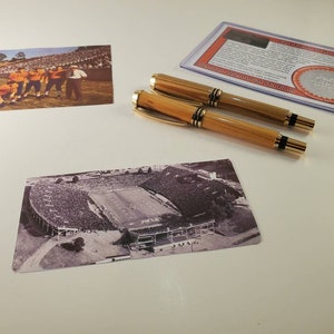May include: Two wooden pens with gold accents are displayed next to two vintage football stadium photographs. One photo shows a team in orange uniforms, and the other is a black and white aerial view of a stadium. A certificate of authenticity is also present.