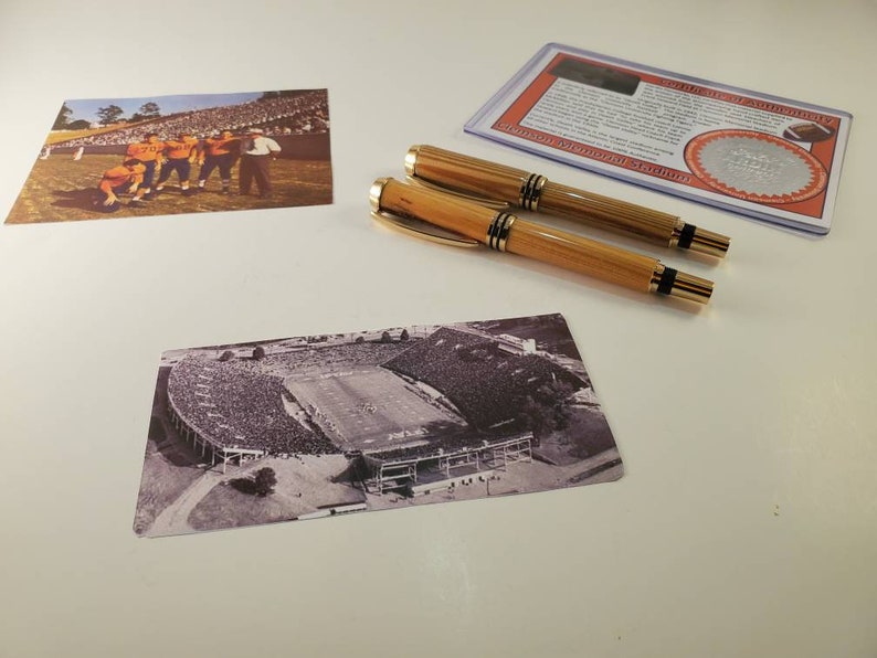 May include: Two wooden pens with gold accents are displayed next to two vintage football stadium photographs. One photo shows a team in orange uniforms, and the other is a black and white aerial view of a stadium. A certificate of authenticity is also present.