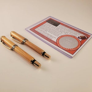 May include: Two wooden pens with gold-colored accents and black tips are displayed next to a certificate of authenticity. The certificate is in a clear plastic sleeve and features text and a football graphic. The pens have a light brown color.