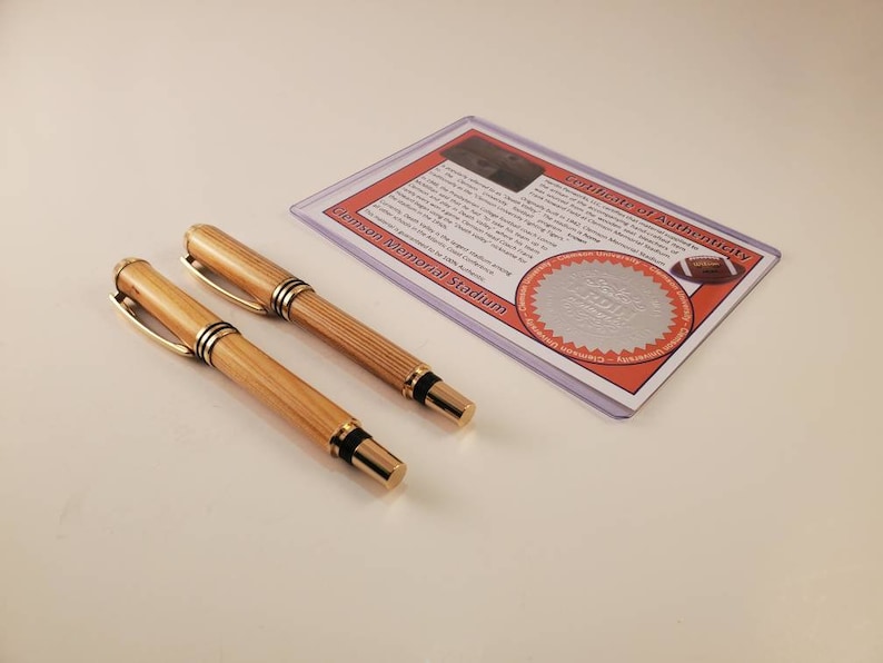 May include: Two wooden pens with gold-colored accents and black tips are displayed next to a certificate of authenticity. The certificate is in a clear plastic sleeve and features text and a football graphic. The pens have a light brown color.