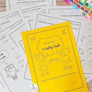 May include: A bright yellow educational activity book titled "أسماء الإشارة هذا وهذه" featuring a line drawing of two children pointing upward, surrounded by several black and white printed worksheets and a collection of colorful markers.