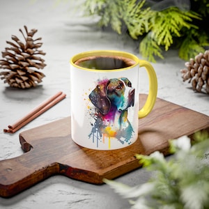 May include: White ceramic mug with a yellow interior and handle, featuring a colorful watercolor painting of a dog's head. The mug is filled with a dark liquid and sits on a wooden cutting board.