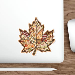 May include: A maple leaf sticker with a patchwork design. The leaf features various patterns, including plaid and floral prints, in shades of orange, red, green, and brown. The sticker has a white border and is placed on a white surface.