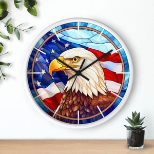 Pode incluir: Relógio de parede branco com um design de vitral de uma águia careca americana com a bandeira americana ao fundo. A águia tem um bico amarelo e penas marrons. O relógio tem ponteiros pretos e algarismos brancos.