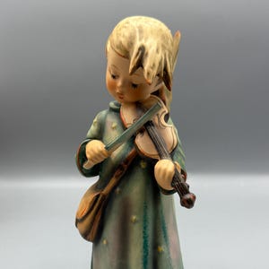 May include: A ceramic figurine of a child playing a violin. The figurine is dressed in a green coat with yellow star accents and a brown satchel. The child has blonde hair and is standing on a square base.