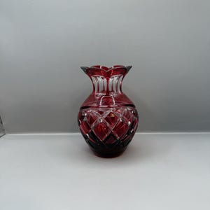 May include: A ruby red glass vase with a diamond-cut pattern. The vase has a flared, scalloped rim and a rounded body. The glass is a deep red color, with a darker shade at the base. A decorative piece for the home.