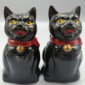 Puede incluir: Par de figuras de gato de cerámica negra con ojos amarillos, pajaritas rojas y detalles dorados. Los gatos tienen un acabado brillante y están sentados. Probablemente sean artículos decorativos.