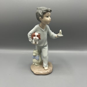 May include: A porcelain figurine of a boy in pyjamas holding a small dog and a plate with a lit candle. The boy is looking at the candle and has a surprised expression on his face. The figurine is standing on a small base.