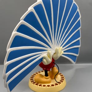 Peut inclure: Figurine d'un personnage de dessin anim&eacute; tenant un &eacute;ventail bleu et blanc. Le personnage porte des chaussures jaunes, un short rouge et des gants blancs. La base est jaune avec une bordure marron.