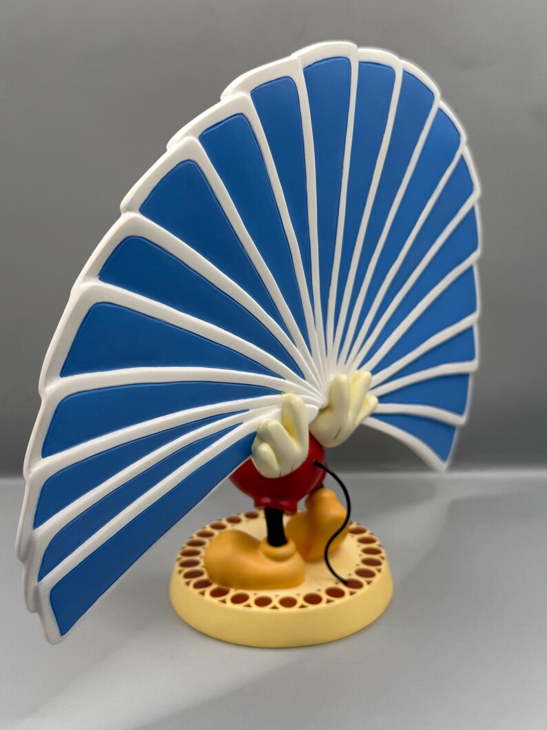 Peut inclure: Figurine d'un personnage de dessin anim&eacute; tenant un &eacute;ventail bleu et blanc. Le personnage porte des chaussures jaunes, un short rouge et des gants blancs. La base est jaune avec une bordure marron.