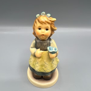 Może przedstawiać: Porcelanowa figurka dziewczynki z brązowymi włosami, ubrana w żółtą sukienkę i trzymająca niebieski kwiat. Ma na głowie niebieską koronę z kwiatów.