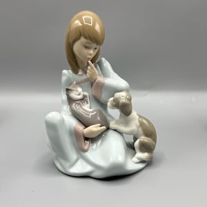 Könnte beinhalten: Porzellanfigur eines sitzenden Mädchens in einem hellblauen Kleid, das eine graue Katze hält und einen kleinen braun-weißen Hund an seiner Seite hat. Das Mädchen hat hellbraunes Haar und berührt mit dem Finger ihre Lippen. Die Figur hat ein glänzendes Finish.