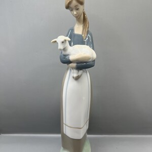 May include: A porcelain figurine of a woman holding a white lamb in her arms. The woman is wearing a blue dress with a white apron and brown shoes. She has brown hair and is looking down at the lamb.