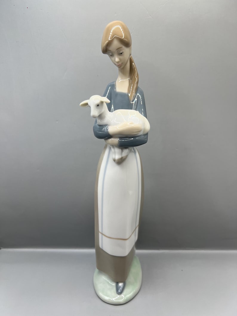 May include: A porcelain figurine of a woman holding a white lamb in her arms. The woman is wearing a blue dress with a white apron and brown shoes. She has brown hair and is looking down at the lamb.