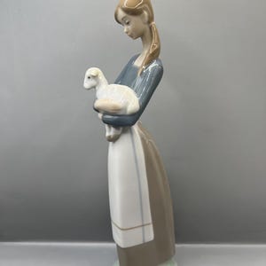 May include: A porcelain figurine of a girl holding a white lamb. The girl is wearing a blue and brown dress with a white apron. The figurine is standing on a green base.