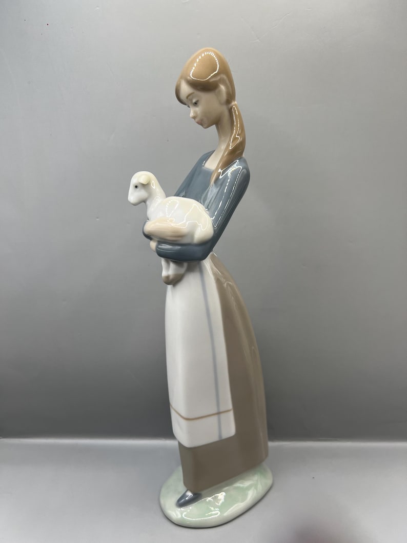 May include: A porcelain figurine of a girl holding a white lamb. The girl is wearing a blue and brown dress with a white apron. The figurine is standing on a green base.