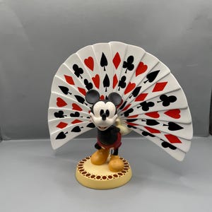 Peut inclure: Figurine de Mickey Mouse tenant un &eacute;ventail de cartes &agrave; jouer. L'&eacute;ventail affiche des enseignes rouges et noires sur fond blanc. Mickey porte sa tenue classique, debout sur une base beige avec une bordure dor&eacute;e. La figurine est pos&eacute;e sur un fond gris.