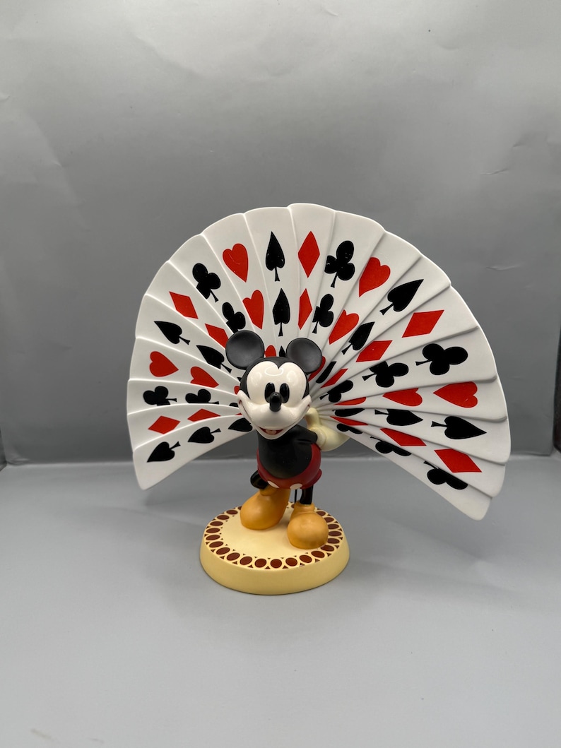 Peut inclure: Figurine de Mickey Mouse tenant un &eacute;ventail de cartes &agrave; jouer. L'&eacute;ventail affiche des enseignes rouges et noires sur fond blanc. Mickey porte sa tenue classique, debout sur une base beige avec une bordure dor&eacute;e. La figurine est pos&eacute;e sur un fond gris.