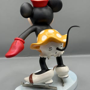 May include: A figurine of a black mouse with large ears, wearing a red hat with a yellow flower, a yellow skirt with white polka dots, and white ice skates. The mouse is in a skating pose on a light blue base.