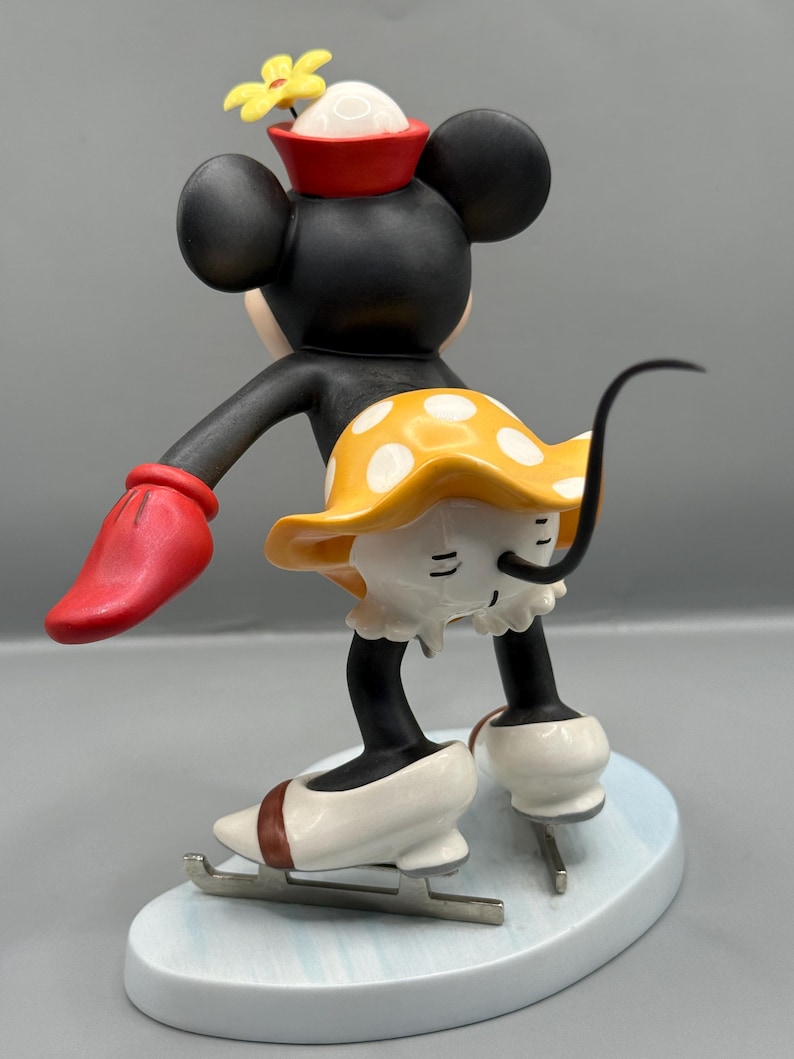 May include: A figurine of a black mouse with large ears, wearing a red hat with a yellow flower, a yellow skirt with white polka dots, and white ice skates. The mouse is in a skating pose on a light blue base.