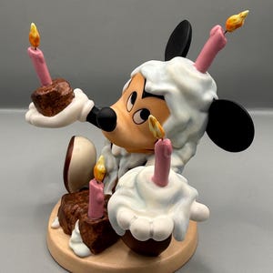 Pu&ograve; includere: Una statuetta stravagante di un topo dei cartoni animati che tiene in mano torte in miniatura con candele accese. Il topo ha una testa bianca e soffice e orecchie nere. Le torte sono marroni con glassa bianca e candele rosa, su una base rotonda marrone chiaro.