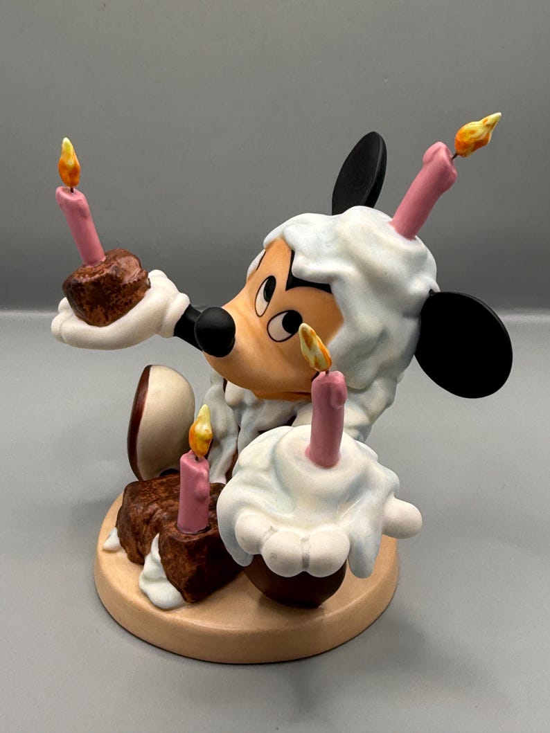 Pu&ograve; includere: Una statuetta stravagante di un topo dei cartoni animati che tiene in mano torte in miniatura con candele accese. Il topo ha una testa bianca e soffice e orecchie nere. Le torte sono marroni con glassa bianca e candele rosa, su una base rotonda marrone chiaro.