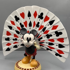 Peut inclure: Figurine de Mickey Mouse avec un &eacute;ventail de cartes &agrave; jouer. La figurine porte un short rouge, une veste noire et des chaussures jaunes. L'&eacute;ventail affiche des enseignes de cartes en rouge et noir. La base est jaune avec une bordure d&eacute;corative.