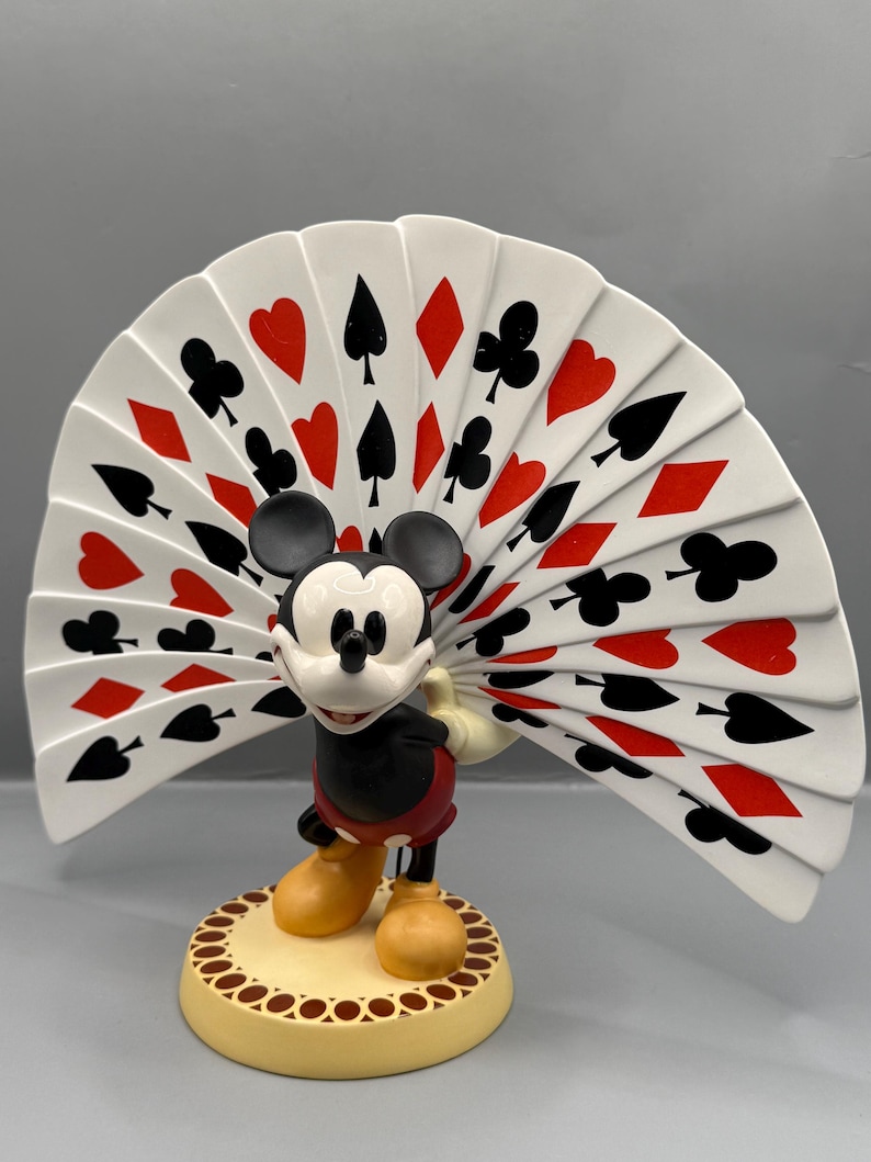Peut inclure: Figurine de Mickey Mouse avec un &eacute;ventail de cartes &agrave; jouer. La figurine porte un short rouge, une veste noire et des chaussures jaunes. L'&eacute;ventail affiche des enseignes de cartes en rouge et noir. La base est jaune avec une bordure d&eacute;corative.