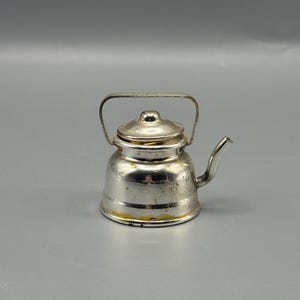 May include: A small, silver-colored teapot with a handle and spout. The teapot has a lid with a small knob on top. The metal has some wear and tear, giving it a vintage appearance. The teapot is likely a decorative item.