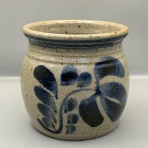 May include: A small, round ceramic pot with a speckled beige base and a dark blue rim. The pot is decorated with a stylized floral design in shades of blue. The pot has a rustic, handmade appearance.