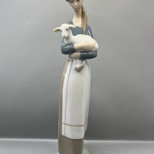 May include: A porcelain figurine of a woman holding a white lamb. The woman is wearing a blue and white dress with a brown apron. The figurine is standing on a green base.