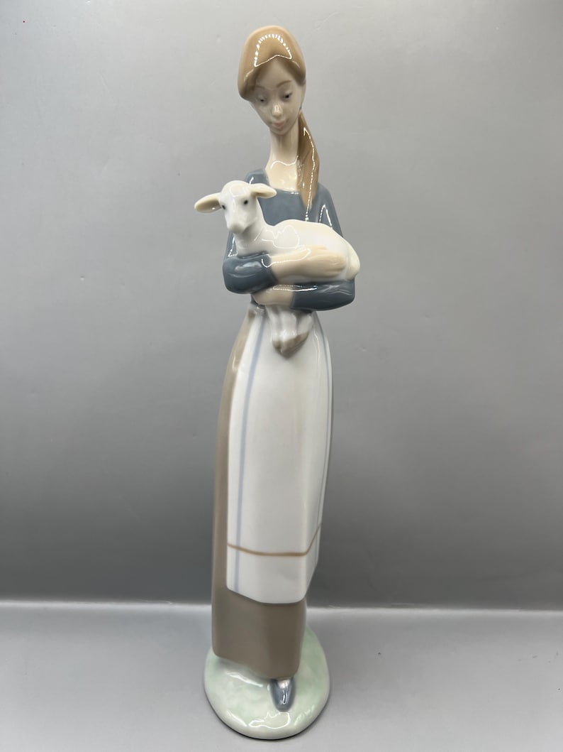 May include: A porcelain figurine of a woman holding a white lamb. The woman is wearing a blue and white dress with a brown apron. The figurine is standing on a green base.