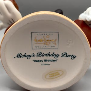 Pu&ograve; includere: Un oggetto da collezione Disney in ceramica, con la festa di compleanno di Topolino. La base &egrave; color crema con la scritta "Mickey's Birthday Party" e "Happy Birthday!". &Egrave; visibile anche il logo Disney Classics Collection.