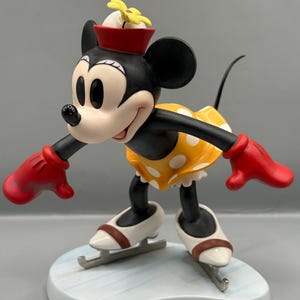 May include: A figurine of Minnie Mouse ice skating. Minnie is wearing a yellow dress with white polka dots, red gloves, and a red hat with a yellow flower. She is on a light blue base, with white ice skates.