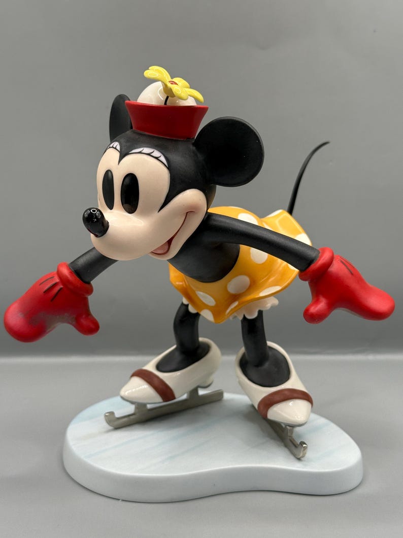 May include: A figurine of Minnie Mouse ice skating. Minnie is wearing a yellow dress with white polka dots, red gloves, and a red hat with a yellow flower. She is on a light blue base, with white ice skates.