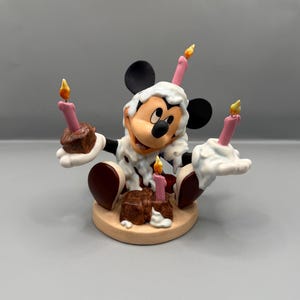 Pu&ograve; includere: Una statuina di Topolino che festeggia un compleanno. La figura indossa una sostanza bianca simile alla glassa e tiene in mano torte al cioccolato con candele rosa. Le candele hanno fiamme gialle. La base &egrave; di colore beige chiaro.