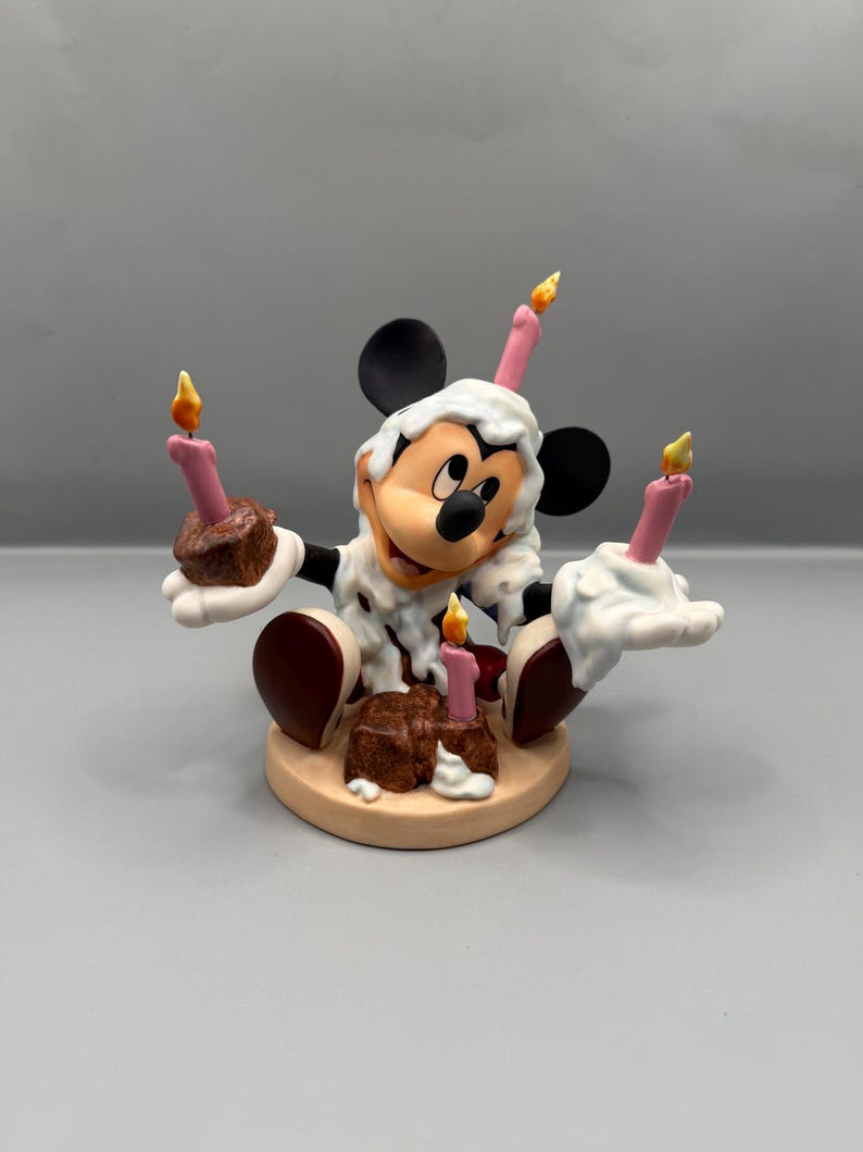 Pu&ograve; includere: Una statuina di Topolino che festeggia un compleanno. La figura indossa una sostanza bianca simile alla glassa e tiene in mano torte al cioccolato con candele rosa. Le candele hanno fiamme gialle. La base &egrave; di colore beige chiaro.