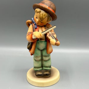 Könnte beinhalten: Eine Figur eines jungen Jungen, der einen braunen Hut trägt und Geige spielt. Er ist in eine braune Jacke, eine grüne Hose und braune Schuhe gekleidet. Die Figur ist aus Porzellan und hat eine bemalte Oberfläche.