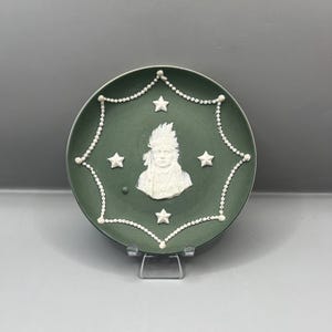 May include: A green ceramic plate with a white relief image of a Native American man wearing a headdress. The plate is decorated with a white beaded border and white stars.