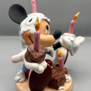 Pu&ograve; includere: Una statuina di Topolino che festeggia un compleanno. La figura ha un cappello di glassa bianca, che tiene in mano due torte al cioccolato con candele rosa. Le candele hanno fiamme gialle. La statuina &egrave; su una base beige.