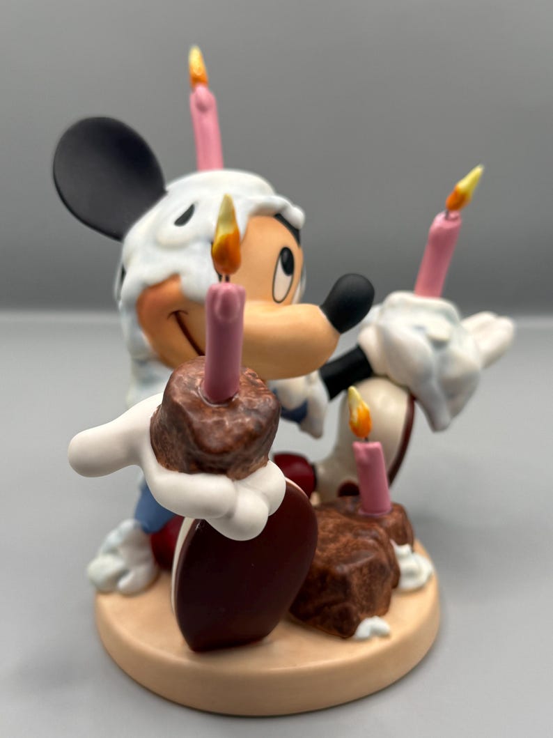 Pu&ograve; includere: Una statuina di Topolino che festeggia un compleanno. La figura ha un cappello di glassa bianca, che tiene in mano due torte al cioccolato con candele rosa. Le candele hanno fiamme gialle. La statuina &egrave; su una base beige.