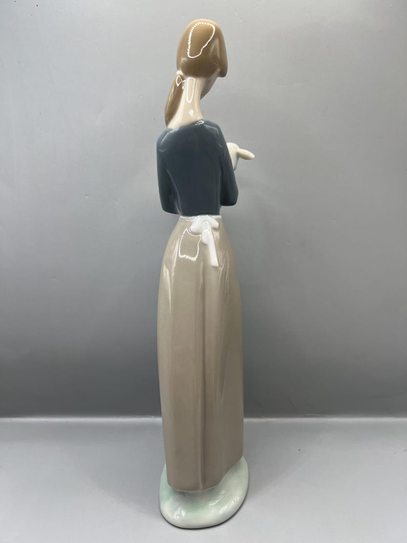 May include: A porcelain figurine of a woman wearing a blue shirt and a brown skirt. She is holding a small white object in her right hand.