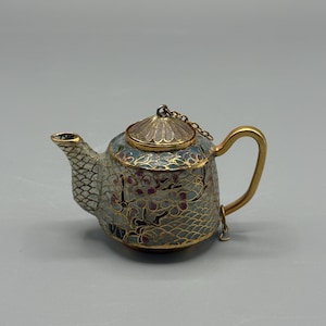 Puede incluir: Una pequeña y ornamentada tetera con asa y boquilla doradas. El cuerpo está decorado con un motivo floral en tonos azules, morados y verdes. La tapa está sujeta con una cadena. La tetera tiene un borde y una base dorados.