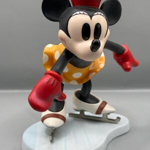 May include: A figurine of Minnie Mouse ice skating. Minnie is wearing a red hat, red gloves, and a yellow dress with white polka dots. Her ice skates are white with brown accents, and she is on a light blue base.