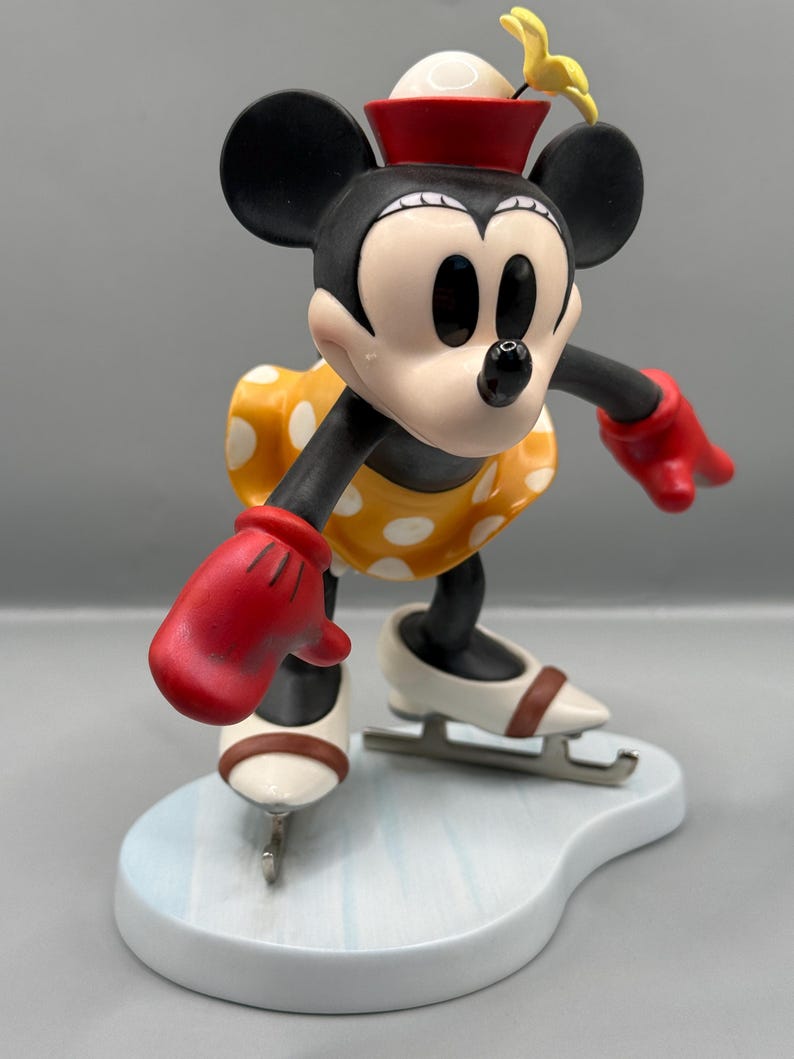 May include: A figurine of Minnie Mouse ice skating. Minnie is wearing a red hat, red gloves, and a yellow dress with white polka dots. Her ice skates are white with brown accents, and she is on a light blue base.