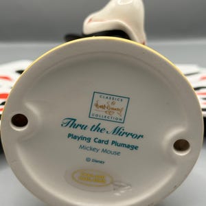 Peut inclure: Figurine en c&eacute;ramique de Mickey Mouse de la collection "Thru the Mirror". La figurine est blanche, noire et jaune, avec le texte "Playing Card Plumage" et "Mickey Mouse". La base a deux trous.