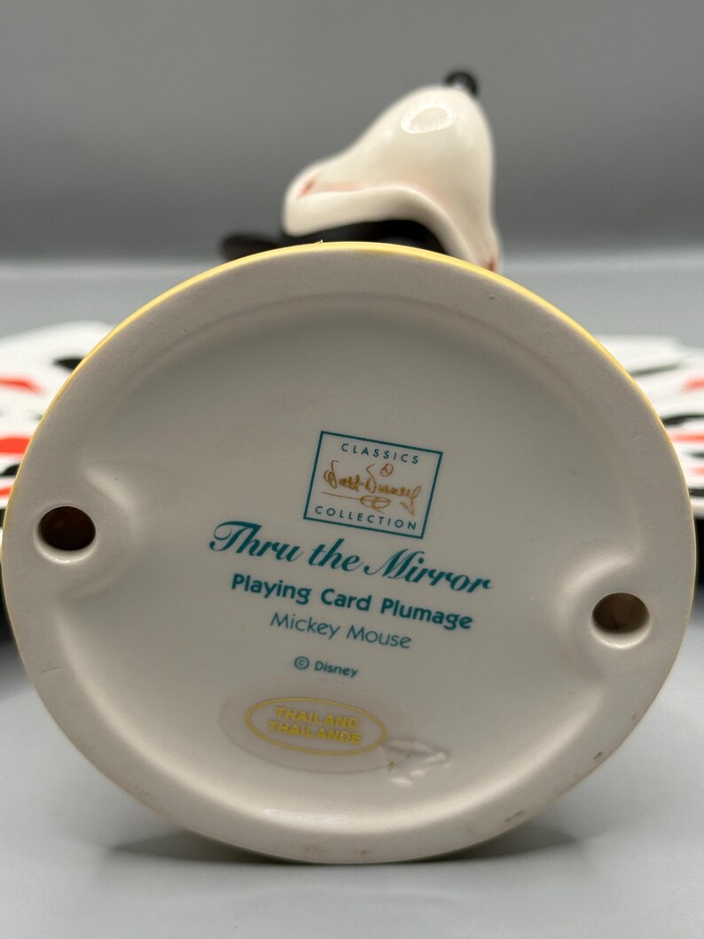 Peut inclure: Figurine en c&eacute;ramique de Mickey Mouse de la collection "Thru the Mirror". La figurine est blanche, noire et jaune, avec le texte "Playing Card Plumage" et "Mickey Mouse". La base a deux trous.