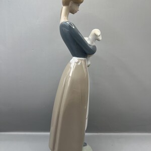 May include: A porcelain figurine of a woman in a blue dress and brown skirt holding a white lamb.